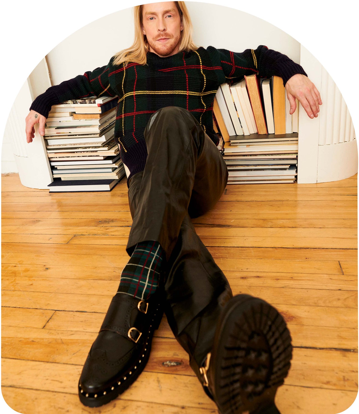 Kate, the double monk derby shoe with studs around the sole, is worn by a male model as he lounges comfortably against some books